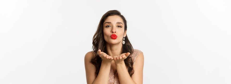 Concept of New Year celebration and winter holidays. Close-up of sensual young woman in dress, pucker lips and blowing air kiss at camera, white background.