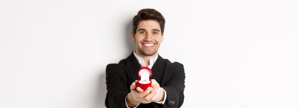 Image of handsome man looking romantic, giving you an engagement ring, making proposal to marry him, standing against white background.