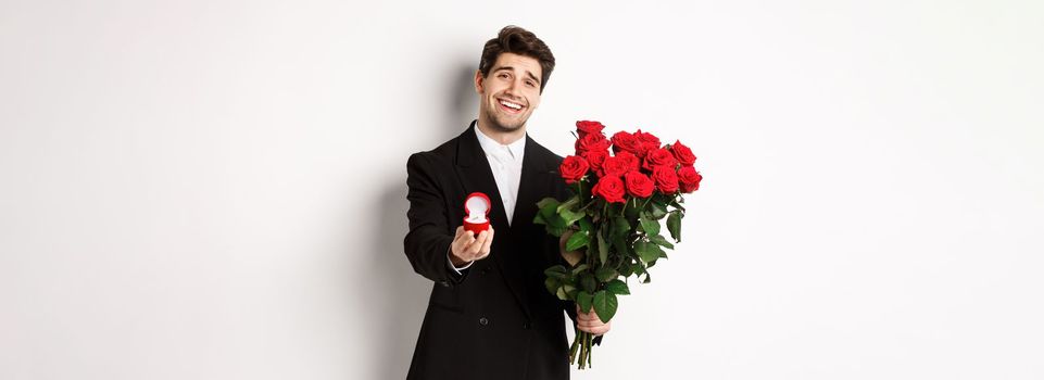 Handsome smiling man in black suit, holding roses and engagement ring, making a proposal to marry him, standing against white background.