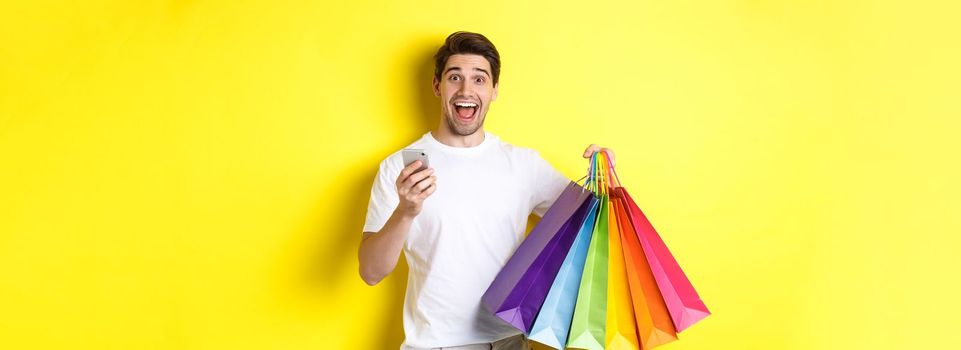 Image of happy man receive cashback for purchase, holding smartphone and shopping bags, smiling excited, standing over yellow background.