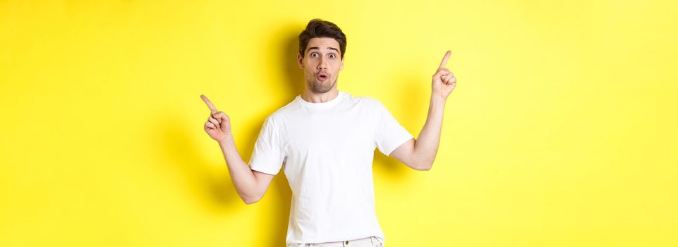 Handsome man pointing fingers sideways, showing two promos, standing over yellow background.