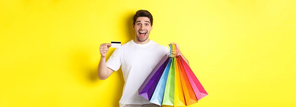 Happy attractive man holding shopping bags, showing credit card, standing over yellow background.