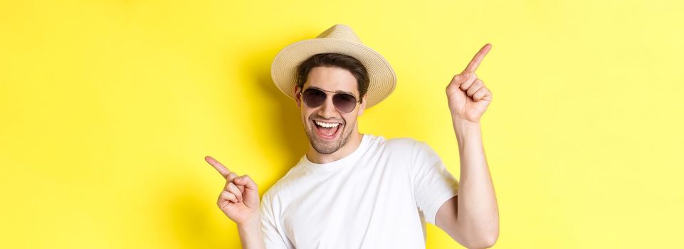 Concept of tourism and vacation. Close-up of man enjoying holidays on trip, dancing and pointing fingers sideways, wearing sunglasses with straw hat.