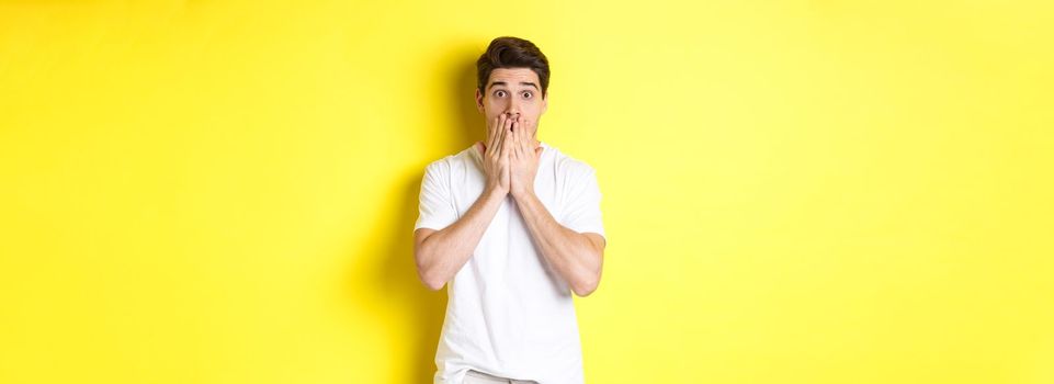 Handsome man looking shocked and speechless, holding hands on mouth, standing over yellow background.
