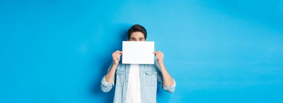 Sneeky handsome guy hiding face behind blank piece of paper for your logo, making announcement or showing promo offer, standing over blue background.