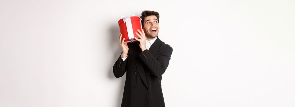 Concept of christmas holidays, celebration and lifestyle. Image of excited man enjoying new year, shaking gift box to guess what inside, standing against white background.