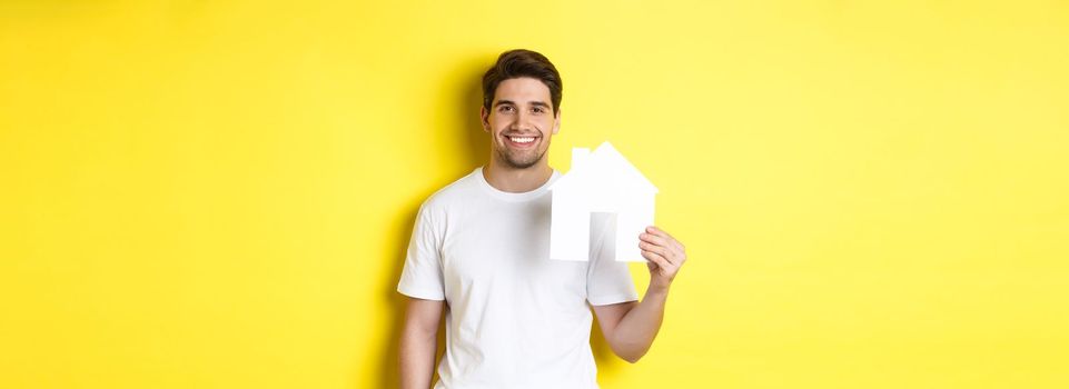 Real estate concept. Young man in white t-shirt holding paper house model and smiling, searching for apartment, standing over yellow background.