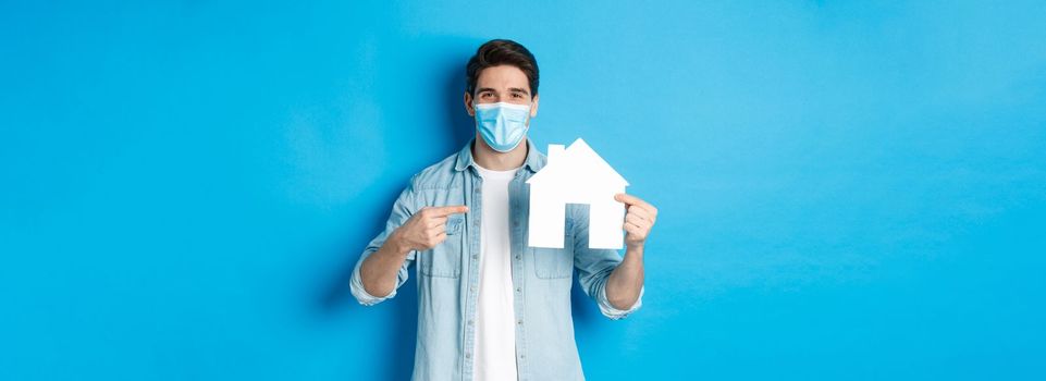 Concept of coronavirus, quarantine and social distancing. Young man searching aparment for rent, business loans, pointing at house model, wearing medical mask, blue background.