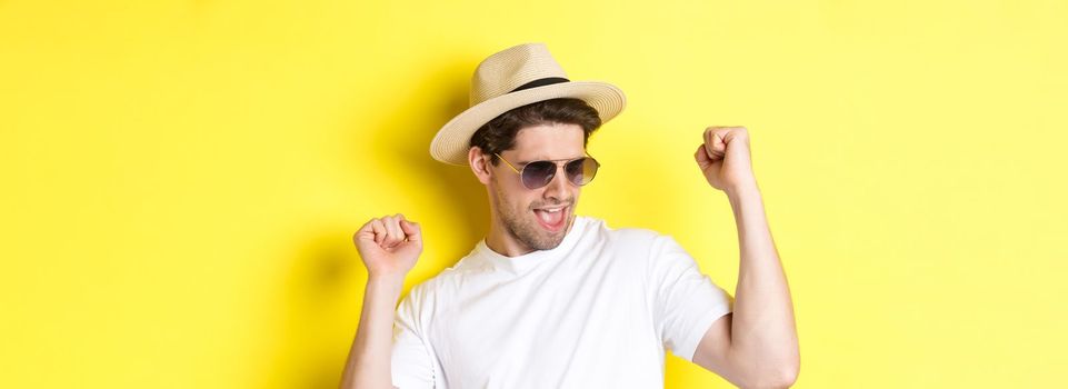 Concept of tourism and vacation. Close-up of man enjoying holidays on trip, dancing and pointing fingers sideways, wearing sunglasses with straw hat, yellow background.