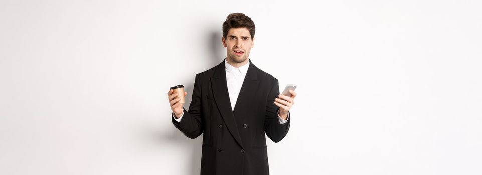 Image of confused businessman standing in a suit, holding cup of coffee and mobile phone, cant understand something, standing against white background.