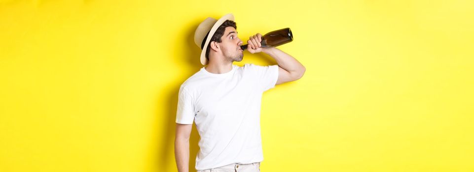 Concept of tourism and vacation. Man drinking wine from bottle on holidays, standing against yellow background. Copy space