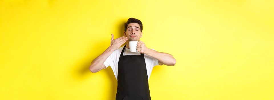Barista enjoying smell of coffee in mug, standing pleased with eyes closed, wearing black apron.