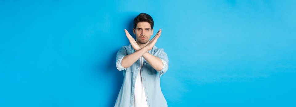 Displeased man frowning and saying no, making cross sign, tell to stop, standing against blue background.