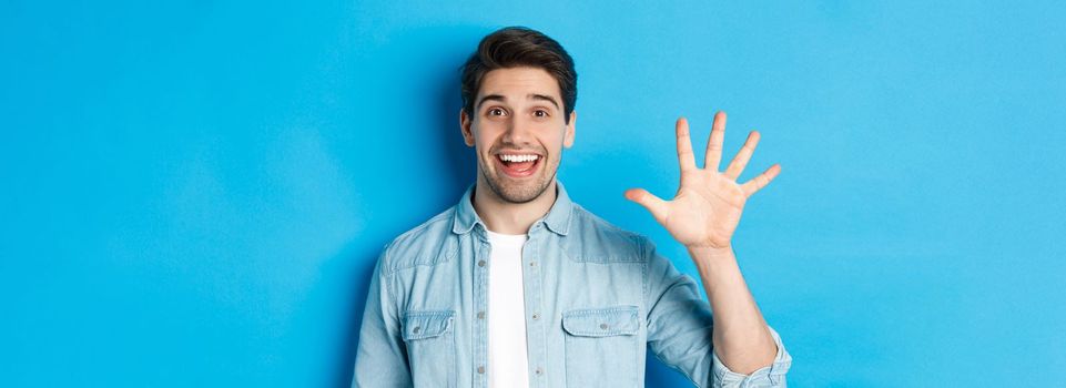 Close-up of handsome man smiling, showing fingers number five, standing over blue background.