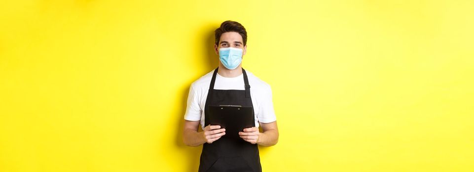 Concept of covid-19, small business and quarantine. Salesman in black apron and medical mask holding clipboard, working in shop, standing over yellow background.
