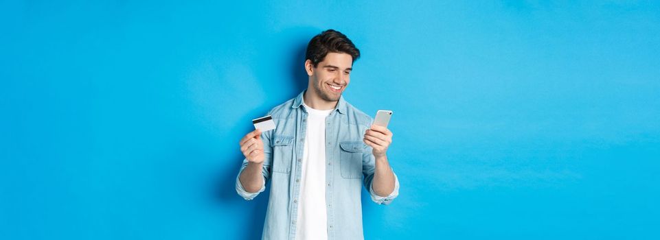 Young man shopping online with mobile application, holding smartphone and credit card, standing over blue background.