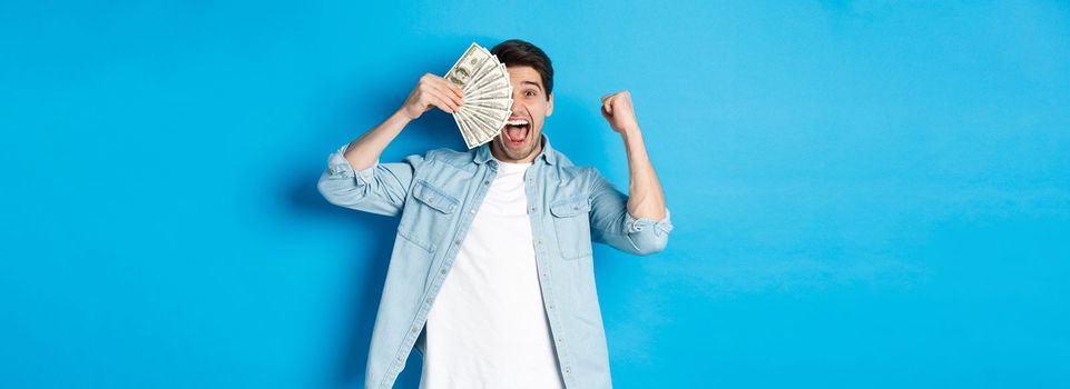 Successful young man earn money, making fist pump and showing cash, winning prize or receive credit, standing over blue background.