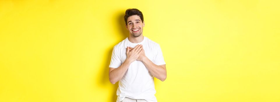 Image of grateful handsome guy in white t-shirt, holding hands on heart and smiling pleased, express gratitude, thanking for something, standing over yellow background.