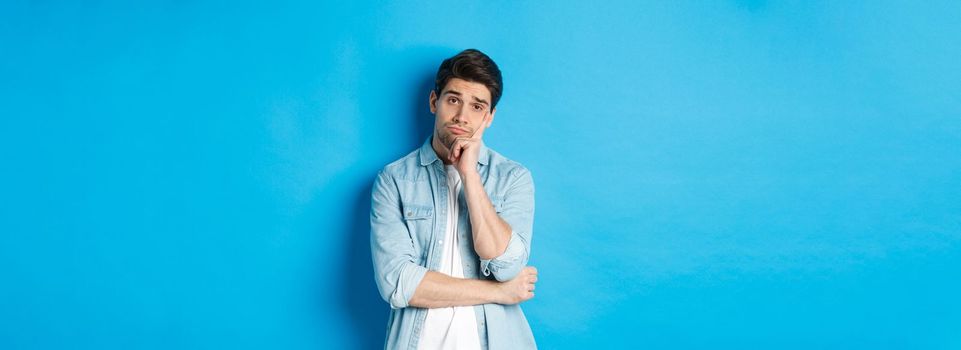 Unamused and bored man looking without interest at camera, standing against bllue background.