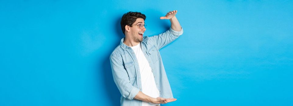 Excited handsome man showing big size object and looking amazed, standing over blue background.