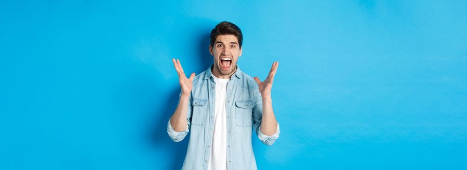 Frustrated young man screaming and looking tensed, shaking hands mad, standing against blue background.