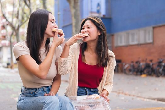 two young women sharing some sweet buns sitting in a city park, concept of friendship and love between people of the same sex