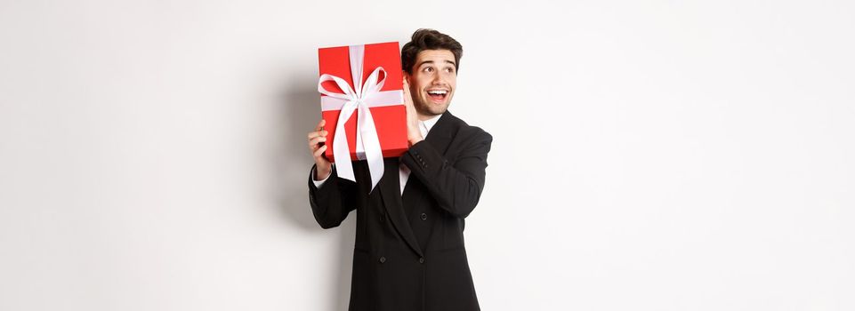 Image of handsome dreamy guy in black suit, shaking box with present to wonder whats inside, standing against white background happy.