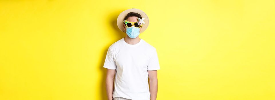 Concept of covid-19, vacation and social distancing. Man tourist wearing medical mask and summer hat with sunglasses, going on trip during pandemic, yellow background.