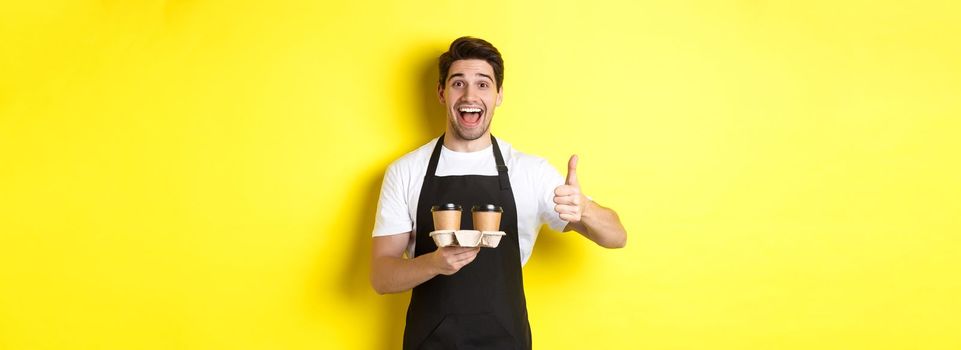 Cheerful barista recommend his cafe, holding takeaway coffee cups and showing thumbs up, standing against yellow background.