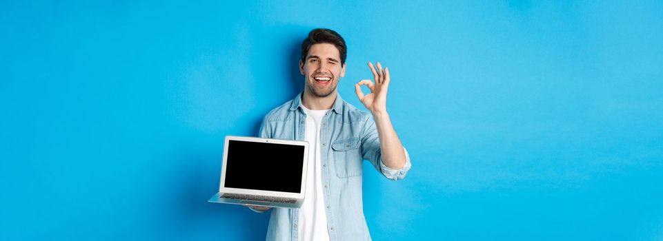 Young man showing laptop screen and okay sign, approve or like promo in internet, smiling satisfied, standing over blue background.