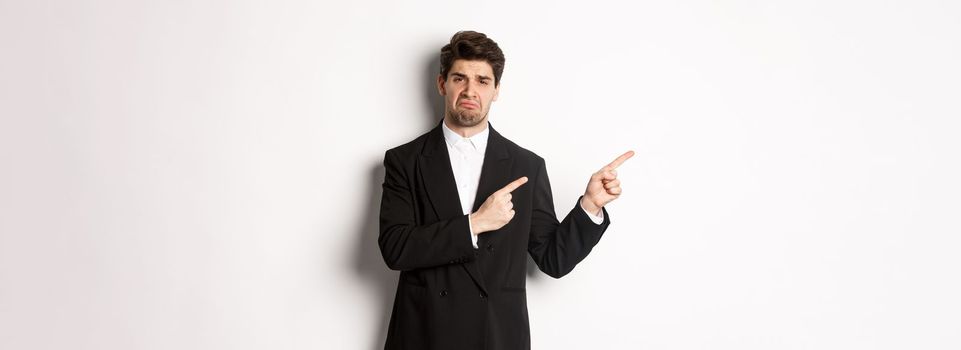 Portrait of disappointed and sad handsome businessman in suit, complaining and pointing fingers left at something bad, standing upset against white background.