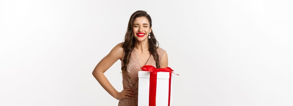 Merry Christmas. Image of attractive woman in luxury dress, celebrating winter holidays, holding gift and laughing from happiness, standing over white background.