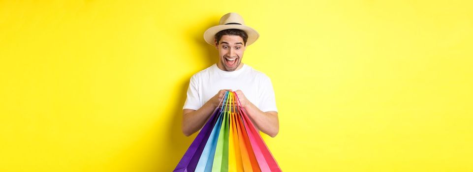 Image of happy man shopping on vacation, holding paper bags and smiling, standing against yellow background.