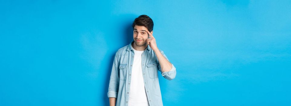 Young thoughtful man pointing at head, asking to think about it, giving a hint, standing in casual clothes over blue background.