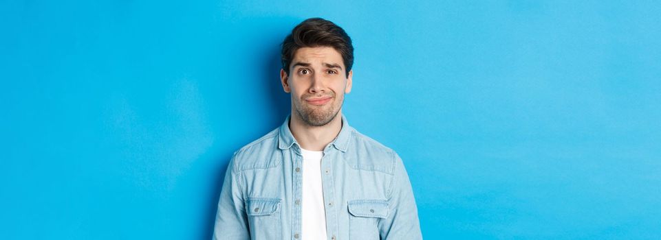 Close-up of skeptical and awkward guy smirking, feeling uncomfortable, standing over blue background.