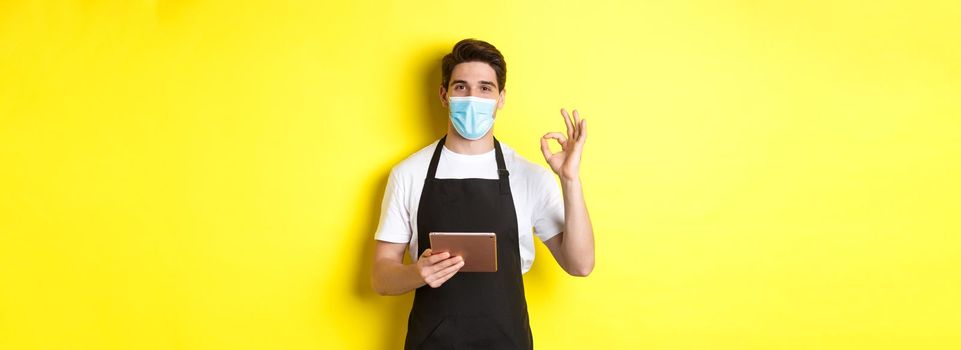 Concept of covid-19, small business and pandemic. Salesman in medical mask and black apron showing ok sign, taking orders with digital tablet, yellow background.