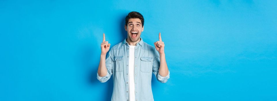 Excited handsome man pointing and looking up, showing advertisement, standing over blue background.