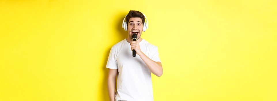 Man in headphones holding microphone, singing karaoke song, standing over yellow background in white clothes.