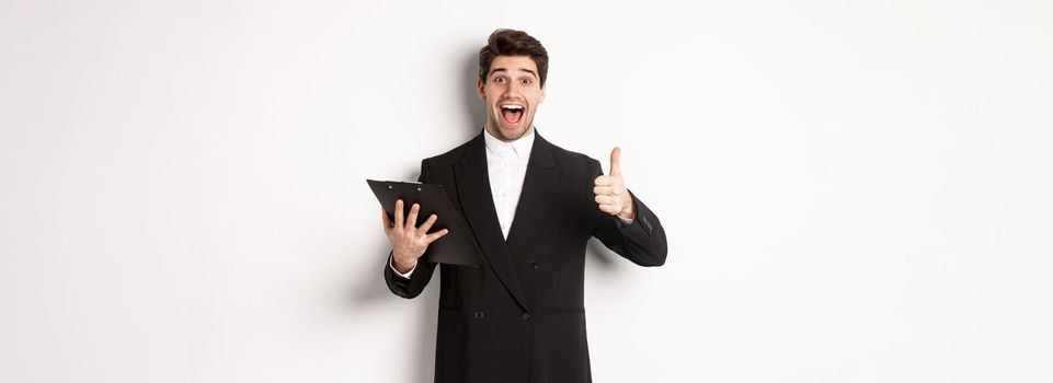 Portrait of confident businessman in black suit, holding clipboard with documents and showing thumb-up in approval, praise good job, standing against white background.