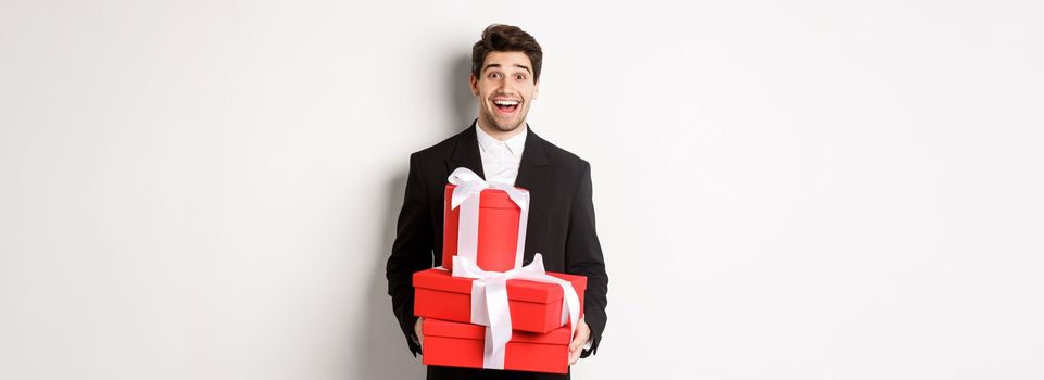 Concept of holidays, relationship and celebration. Handsome man in black suit bringing presents at new year party, holding gifts and smiling amused, standing against white background.