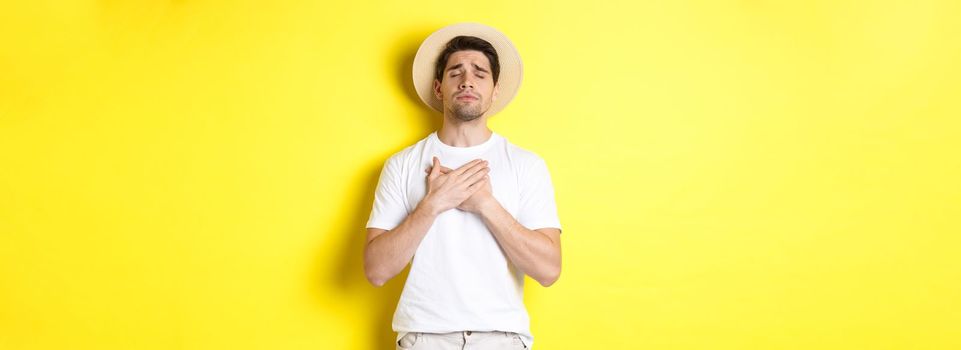 Concept of tourism and summer. Romantic man in straw hat looking nostalgic, close eyes and holding hands on heart, standing against yellow background.