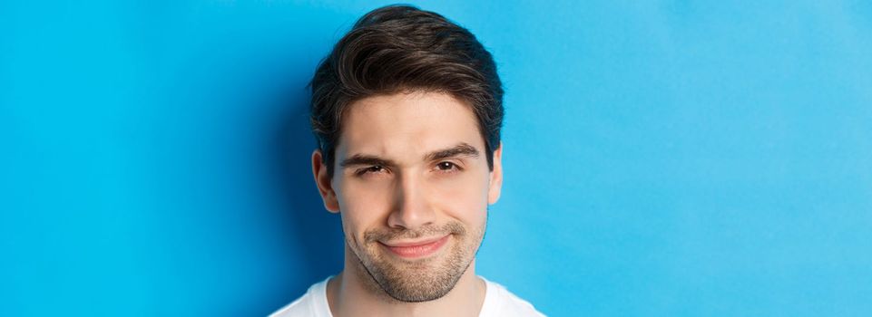 Headshot of thoughtful handsome man, looking intrigued and squinting, thinking about something, standing over blue background.