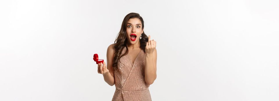 Excited young woman in glamour dress being engaged, showing finger with ring and looking happy, receive marriage proposal, standing over white background.