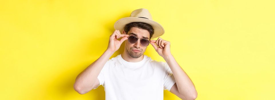 Concept of tourism and vacation. Close-up of cool tourist enjoying holidays on trip, wearing sunglasses with straw hat, yellow background.