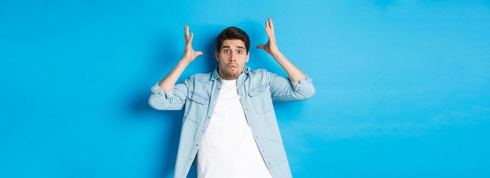 Distressed guy showing mind blowing gesture, looking frustrated and anxious, standing against blue background.