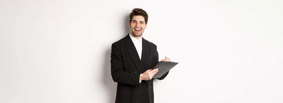 Image of handsome, successful boss in black suit, winking and smiling while signing documents, standing against white background.