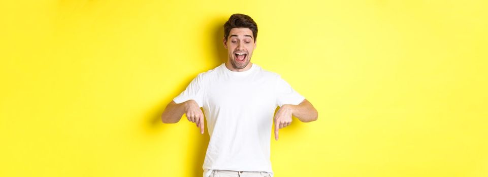 Image of amused handsome guy in white t-shirt, looking and pointing fingers down, standing over yellow background.