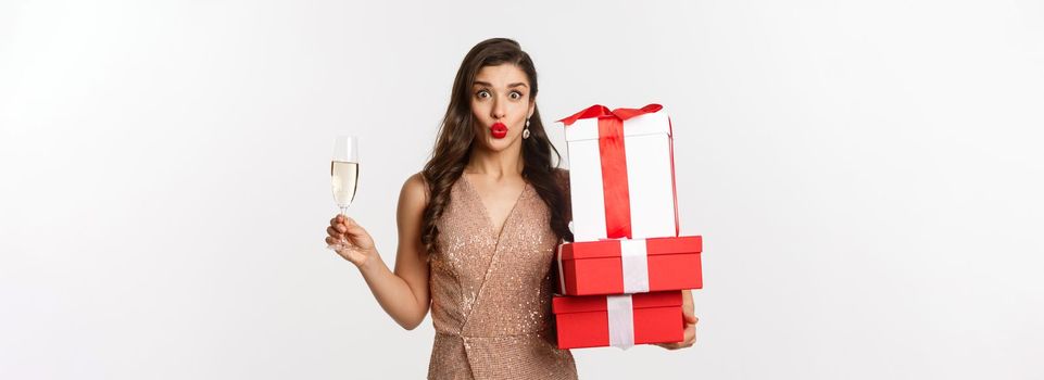 New Year, Christmas and celebration concept. Elegant woman in luxury dress and red lipstick, holding gifts and drinking champagne on party, standing over white background.