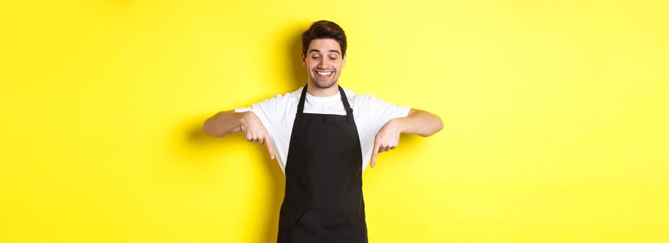 Happy coffee shop worker in black apron pointing fingers down, looking pleased and smiling, showing logo, standing over yellow background.