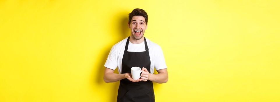 Happy barista in black apron holding coffee cup, laughing and standing over yellow background.